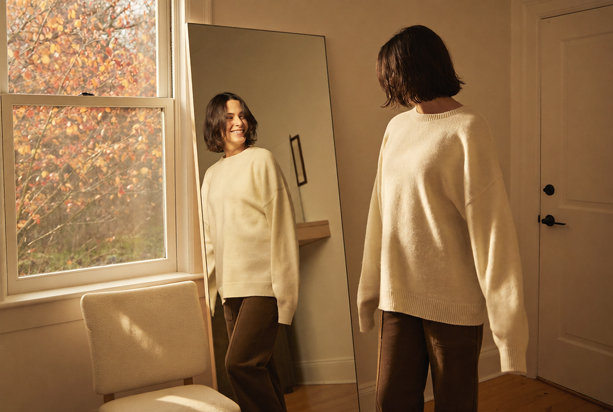 Woman trying on a cream knit sweater at home in front of a mirror